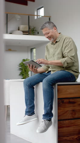 vertical video of happy senior biracial man sitting on table using tablet at home, slow motion