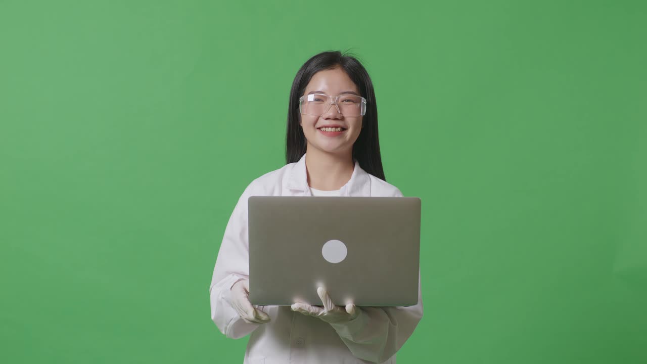 아시아 여성 과학자가 녹색 화면 배경 실험실에서 노트북에 서서 작업하는 동안 카메라에 미소 짓고 있습니다.