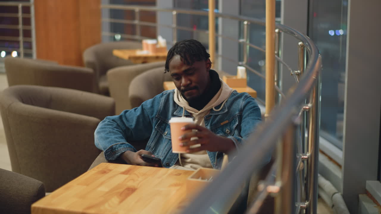 Young man in denim jacket and beige hoodie sits relaxed in cozy modern cafe at night, holding smartphone and takeaway coffee cup while gazing down thoughtfully, surrounded by soft lighting and warm tones