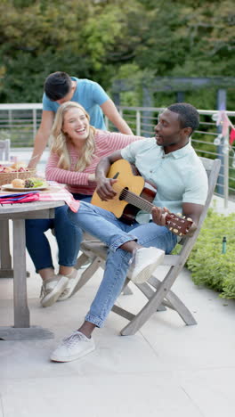 vertical video of african american man playing guitar with diverse friends in garden, slow motion