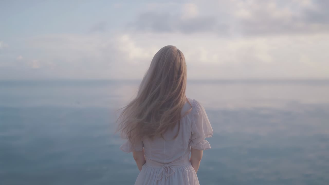 Feeling breeze, woman standing at water edge with hair and dress flowing admiring calm sea