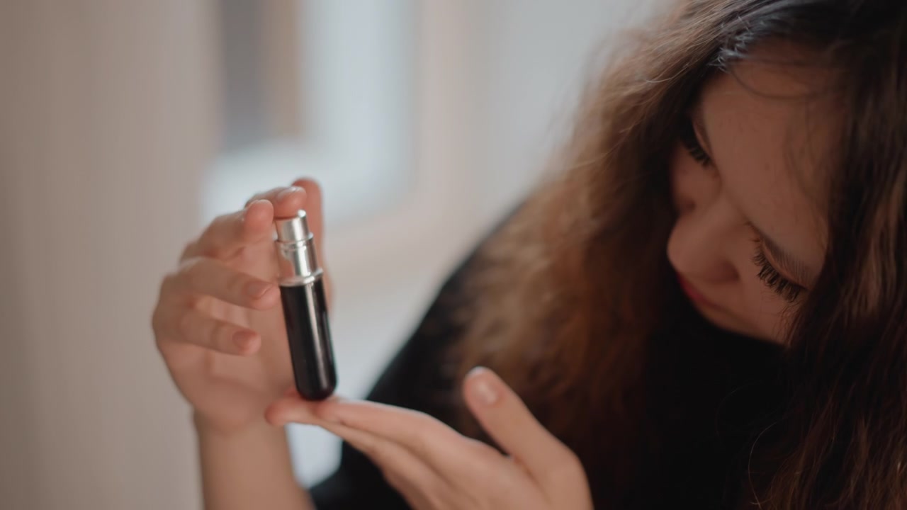 Long curly-haired young woman tilts head and dispenses liquid from small dropper bottle onto fingertip, showing careful hand movement, focused attention, soft indoor light, and natural beauty routine