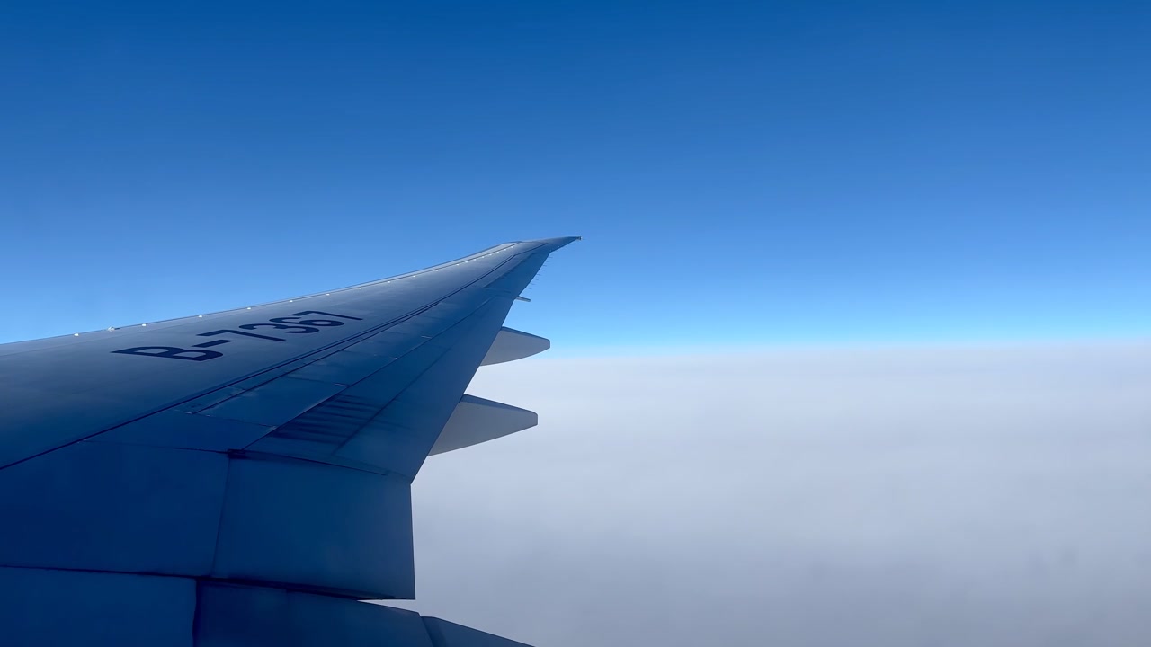 Vista dal finestrino di un aereo che mostra l'ala sopra un vasto strato di nuvole bianche. Un cielo azzurro limpido si estende lungo l'orizzonte durante il volo sopra la Russia