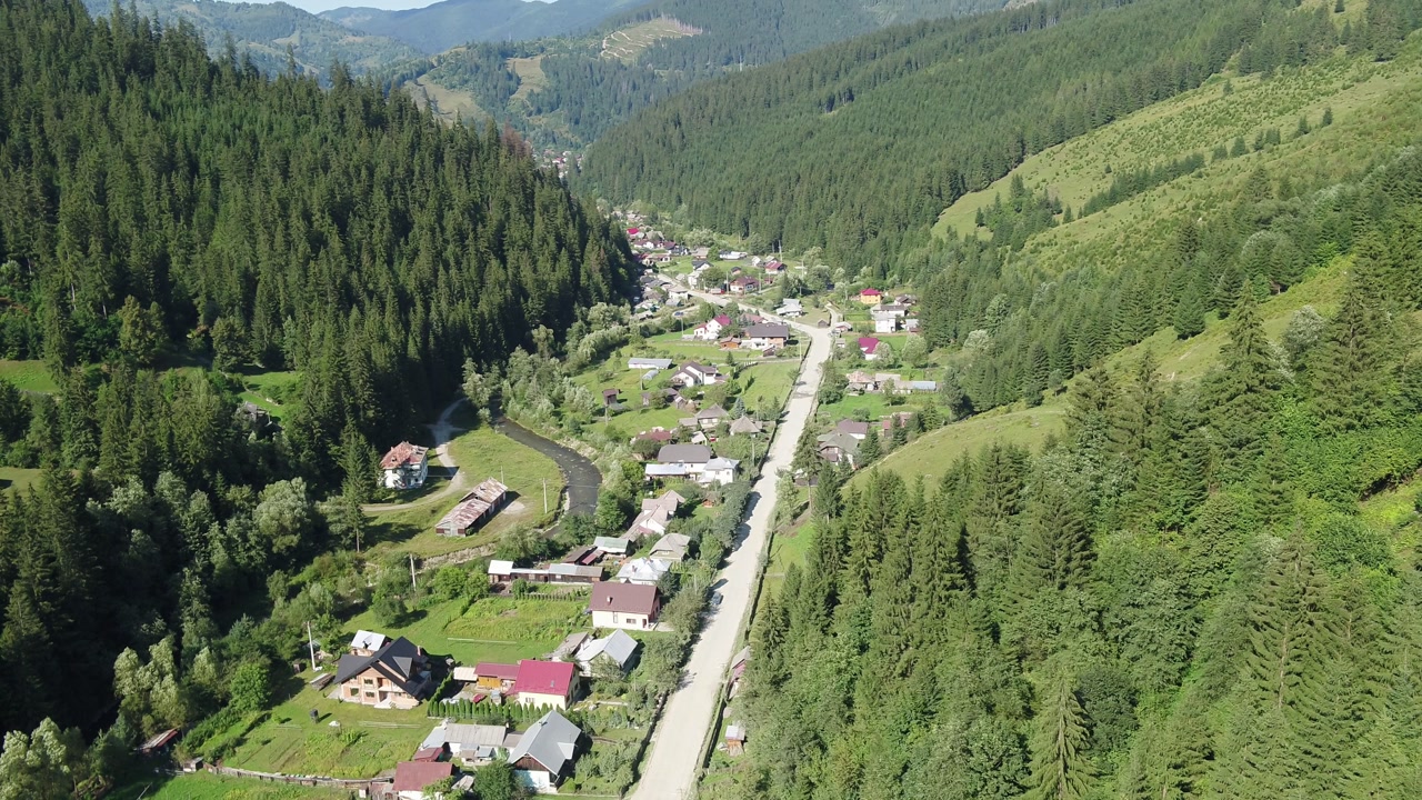 A high-angle aerial shot slowly orbits over a rural village in a Romanian mountain valley. Houses are scattered along a river and road, surrounded by lush green summer forests. Brosteni, Suceava.