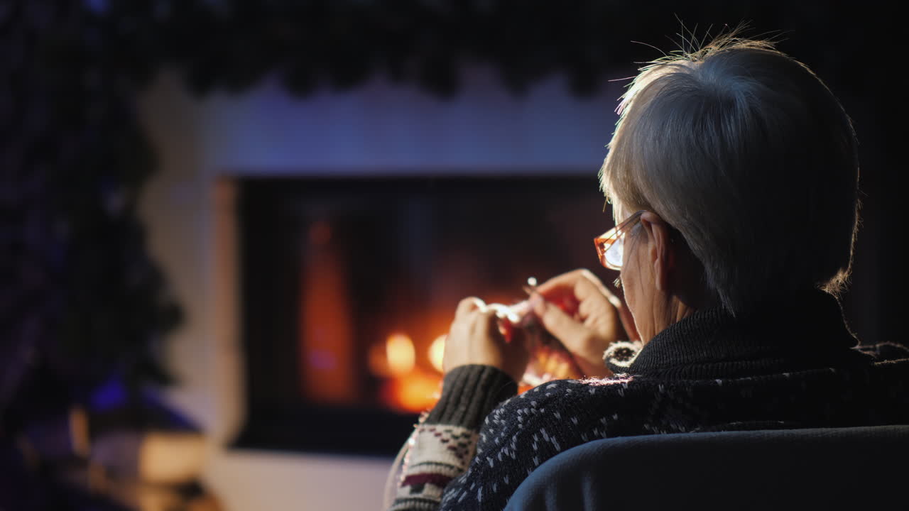 크리스마스 이브에 벽난로 옆에서 저녁에 한 할머니가 따뜻한 옷을 짠다 4k 비디오