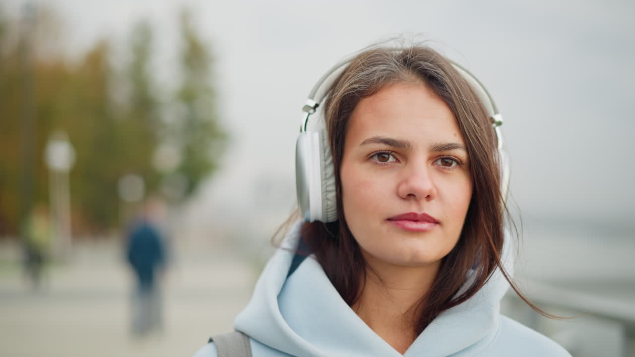 Close-up view of woman listening to music with headphones, eyes closed, serene expression, with blurred view of two people walking behind her in peaceful outdoor urban setting