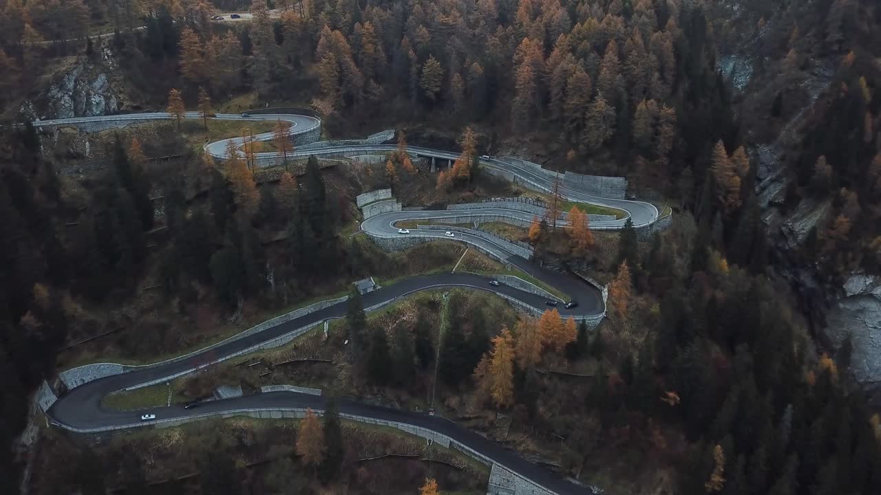 Aerial view of a winding mountain road with multiple hairpin turns, surrounded by autumn-colored trees and rugged terrain, showcasing a scenic and challenging route through a forested landscape