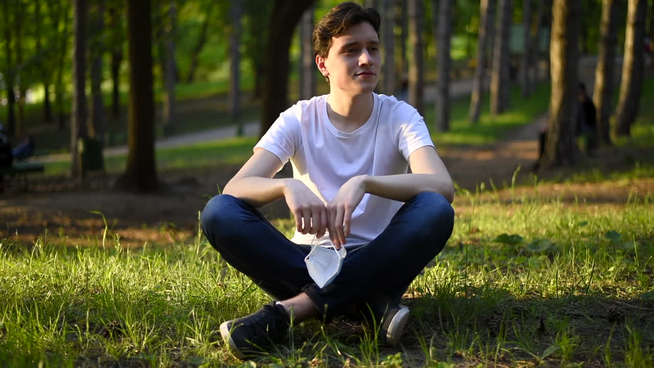 Young man in white t-shirt taking off a protective medical mask from the face and inspire air while resting on grass in the park. Sunny day. Corona Virus idea