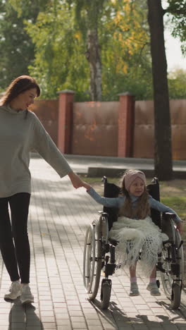 woman walks on stone road holding hand of little daughter with muscular dystrophy. caring mother spends time in park with little girl in wheelchair