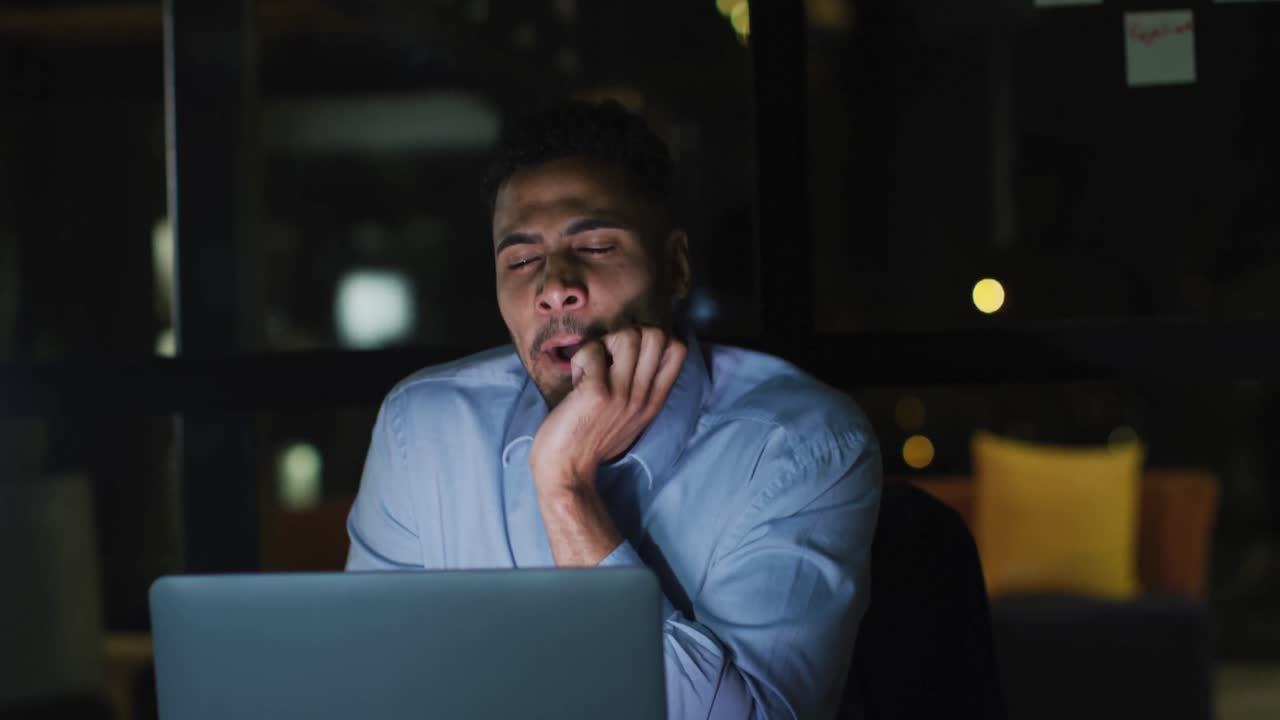 Video of tired biracial businessman sitting at desk using laptop, working at night in office