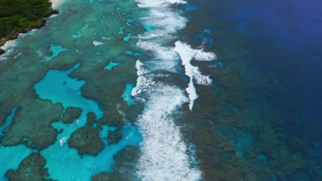 a drone captures a shot of a blue lagoon reef with waves crashing, displaying the vibrant colors and movement of the water, panning up to reveal the stunning coastal landscape of a tropical island