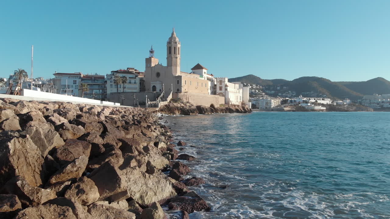 el muelle rocoso se extiende hasta una antigua iglesia construida en piedra, asentada al borde del mar en medio de olas rompientes y rodeada de imponentes montañas