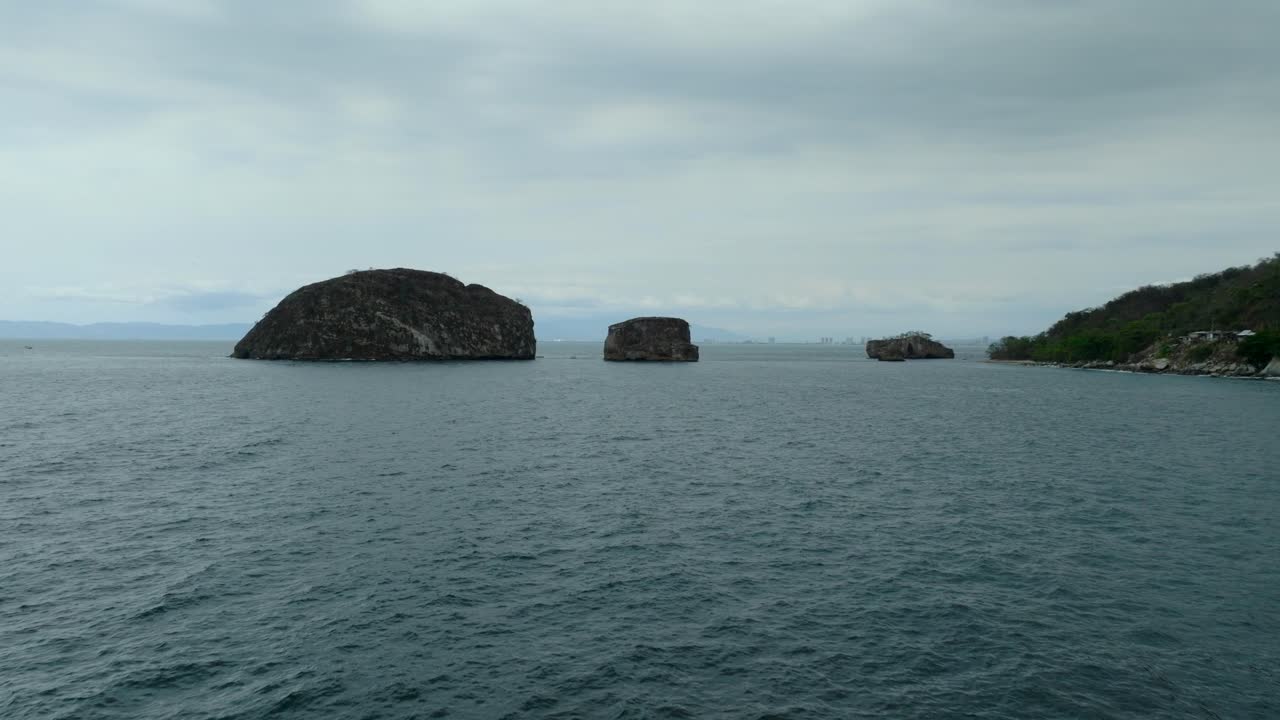 Low angle aerial flight over the Pacific ocean toward Los Arcos de Mismaloya rocky islands near Puerto Vallarta, Mexico. Overcast sky