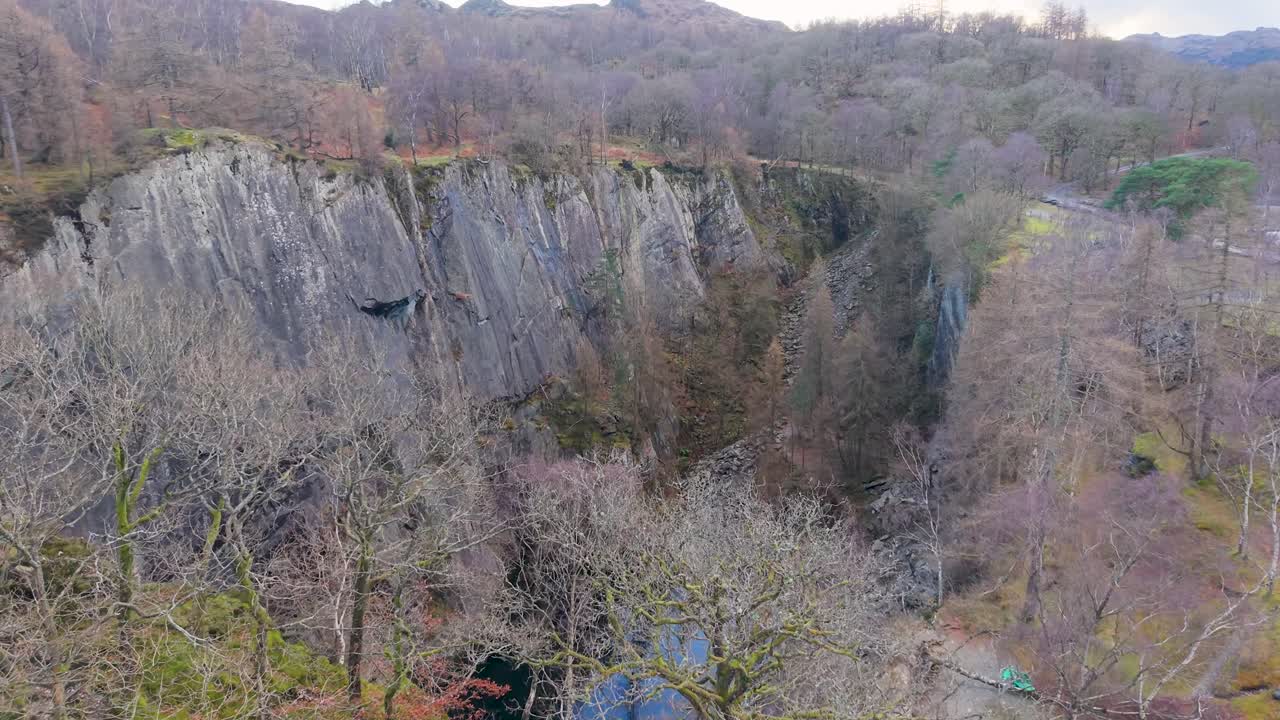A drone flies toward a deep quarry, surrounded by trees in winter, tilting downward to reveal dramatic rock walls and a still pool reflecting the sky and clouds in this striking aerial scene