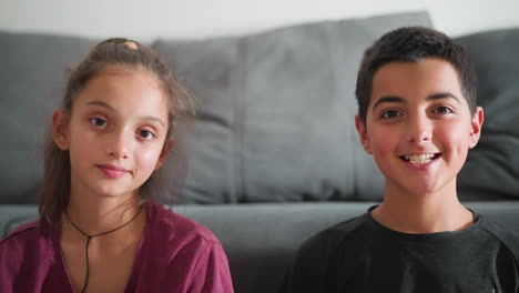 portrait view of beautiful friends sitting indoors looking directly at camera with boy smiling joyfully and blinking eyes while girl calmly gazes creating warm relaxed atmosphere with soft lighting