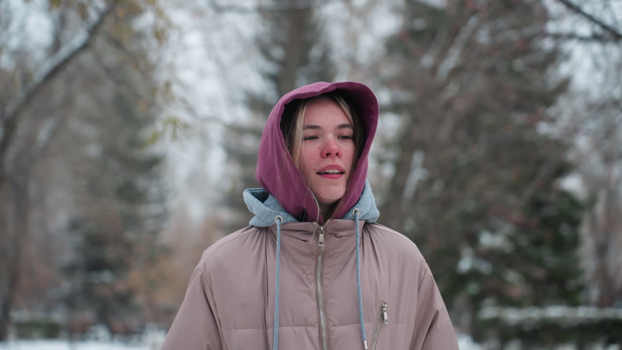 추운 겨울 공원에서 조하는 여인, 사려 깊은 표정으로, 따뜻한 후디와 코트를 입고, 배경에는 눈으로 인 나무