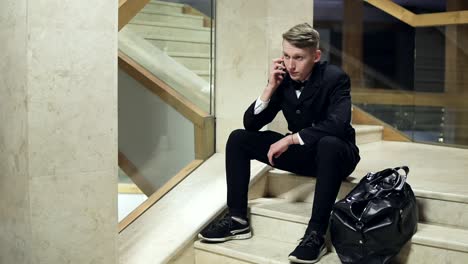 young blonde man in black suit with big black leather bag dialing a number but no one answering. walking away