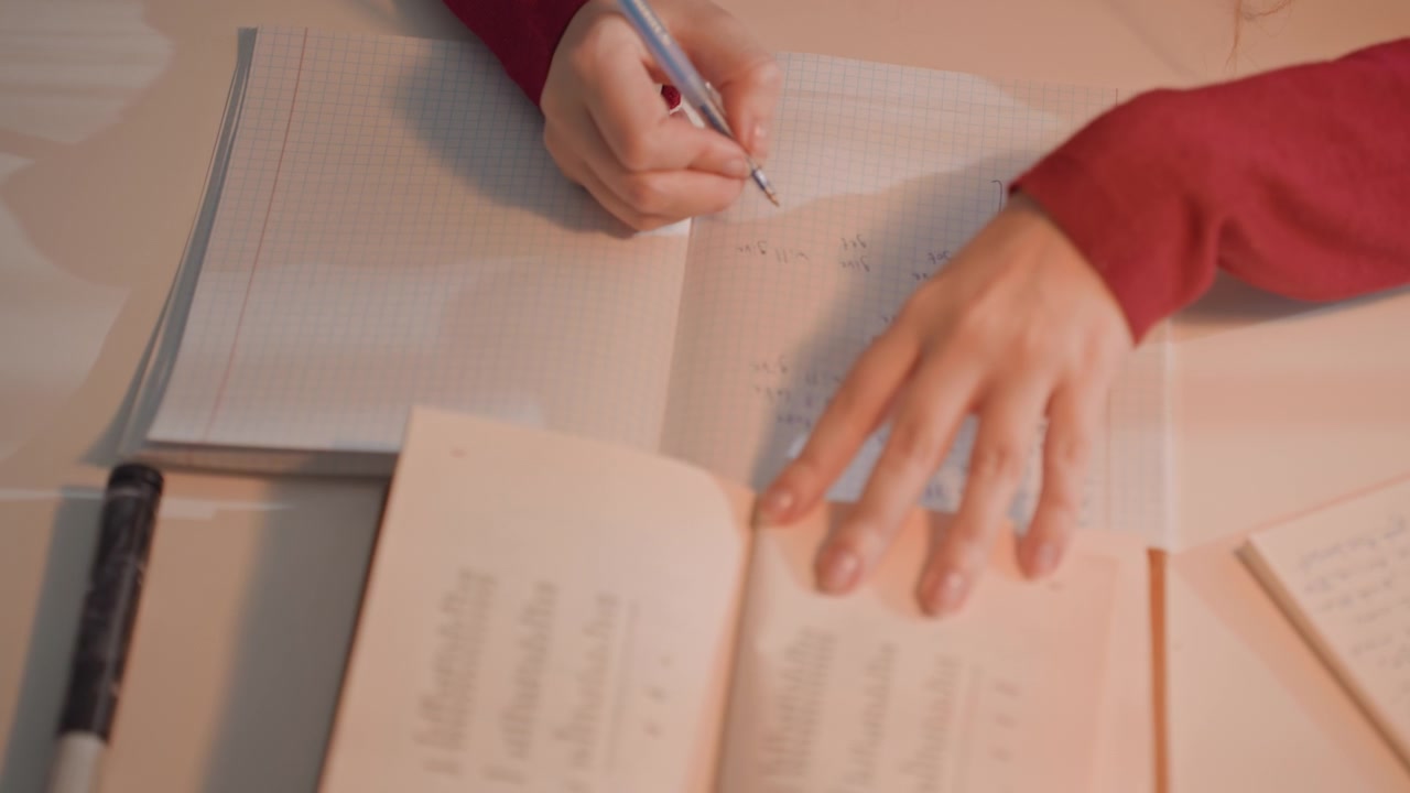 Person i rødt langermet plagg skriver matematiske ligninger. Skrivebordet inkluderer åpne bøker og flere fylte notatbøker. Myk belysning lyser opp arbeidsplassen og fremhever studiet.