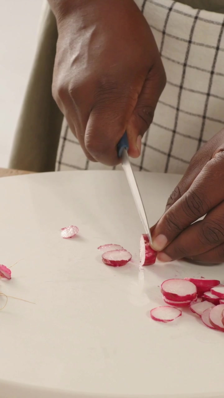 Vertical video: Placing radish, using blue knife, aproned cook hands slicing rounds on white board