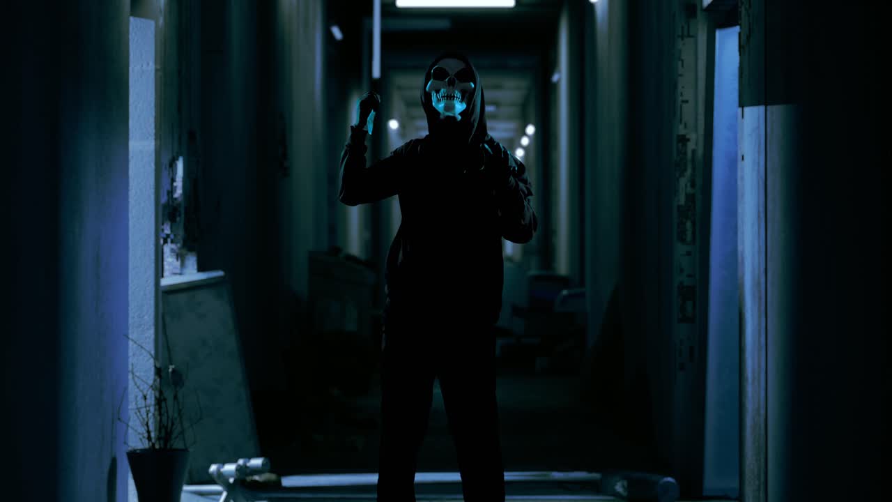 hombre aterrador con capucha usando máscara de fantasma apuñalando con un cuchillo por hacer un gesto amenazante mientras sostenía un cuchillos en el hotel abandonado, escena de terror