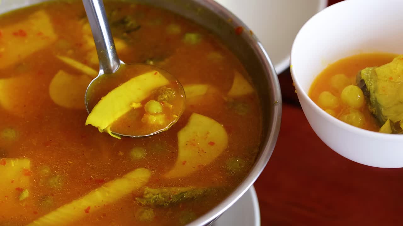 A ladle serves vibrant, spicy soup with vegetables into a white bowl on a wooden table.