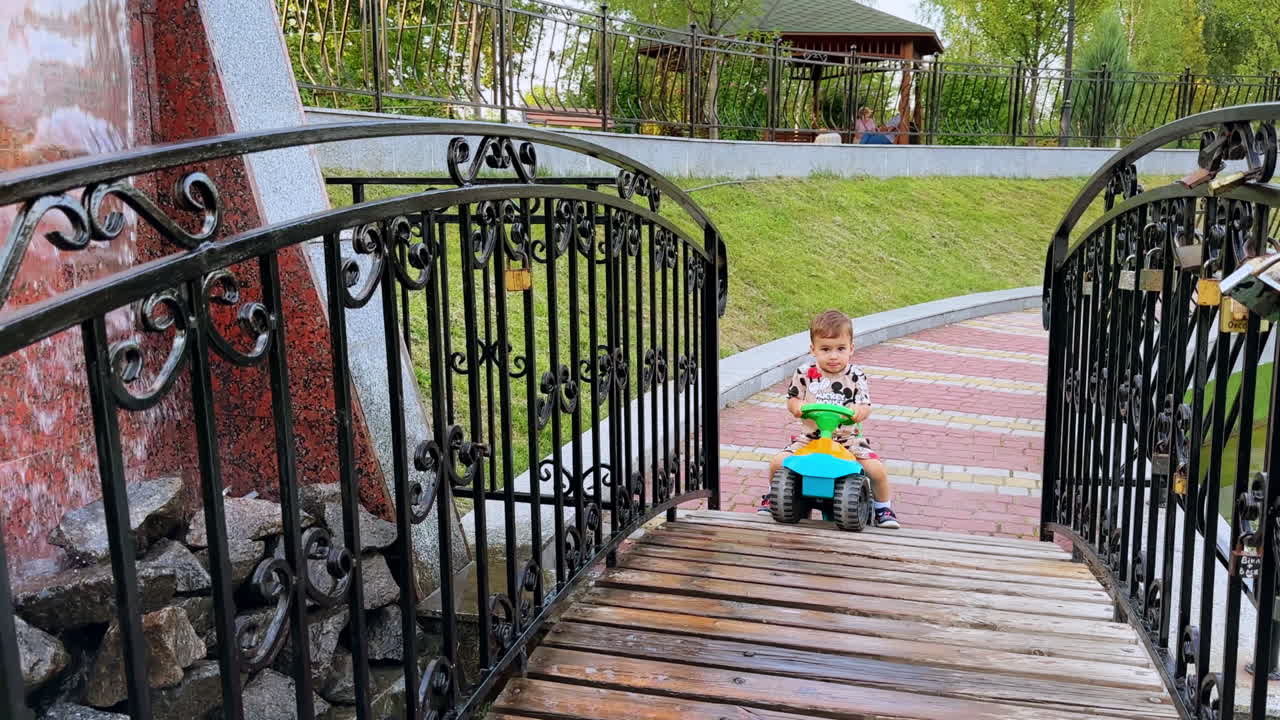 Adorable toddler sitting on the toy car tries to get onto the wooden bridge. Little boy on the walk in the park in summer.
