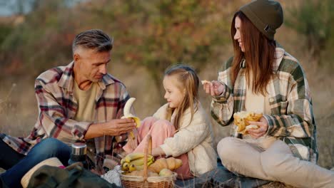 A-brunette-man-in-a-Green-checkered-shirt,-together-with-his-wife,-a-brunette-girl-in-a-Green-checkered-shirt-and-their-little-daughter,-are-sitting-outside-the-city-on-a-picnic-and-eating-fruits-in-the-summer-on-vacation