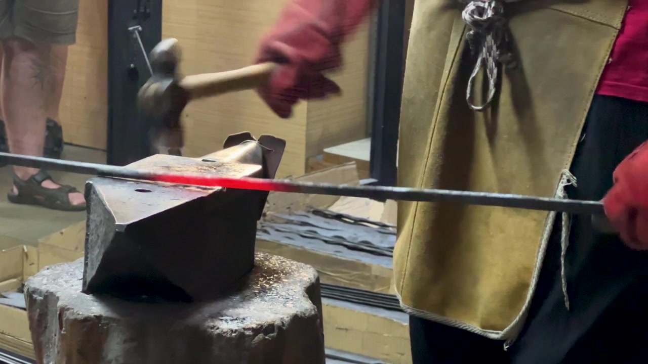A skilled blacksmith in protective gear hammers a glowing red hot metal rod on a traditional anvil. Authentic craftsmanship and manual labor in a metalworking studio.