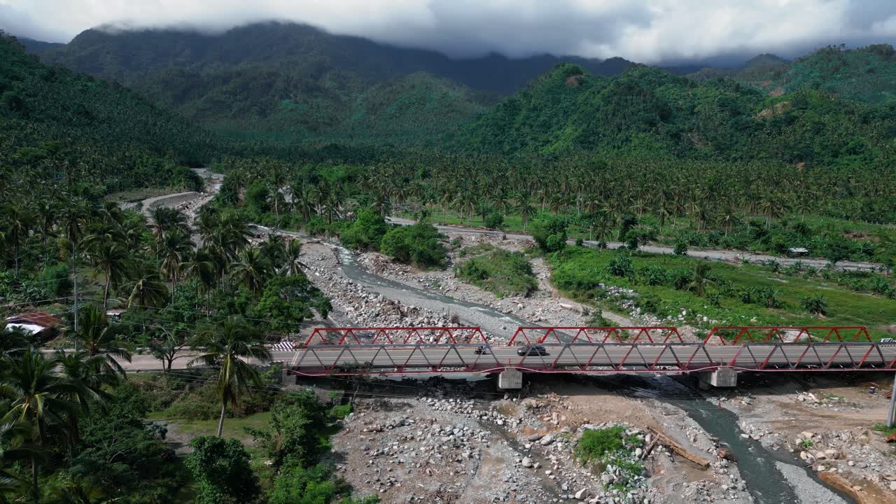 A wide shot captures the majestic mountains, with fluffy clouds drifting across the sky. Palm trees line the foreground, and a bridge stretches across the landscape, creating a stunning view of nature