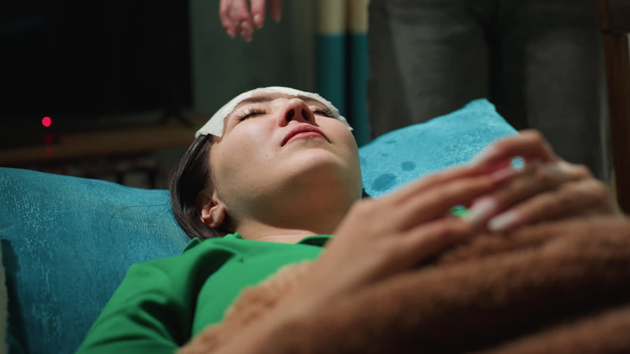 Una mujer asiática tumbada en un sofá recibe una suave compresión facial de una esteticista en un spa tenue y acogedor, toalla turquesa y manta marrón, manos en primer plano aplican una almohadilla fría en la frente, respiración lenta.