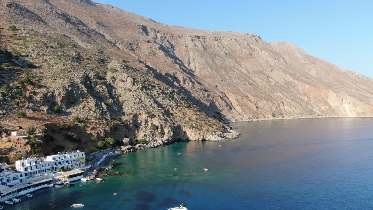 Drone view in Greece flying over blue sea in Loutro small white house town and small boats next to a hill on a sunny day