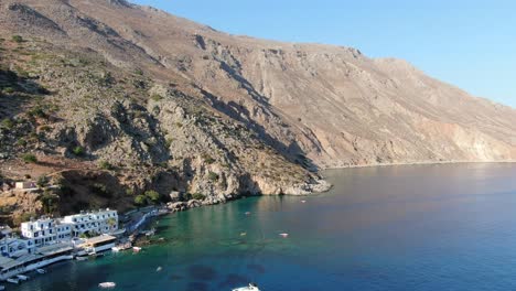 Drone-view-in-Greece-flying-over-blue-sea-in-Loutro-small-white-house-town-and-small-boats-next-to-a-hill-on-a-sunny-day