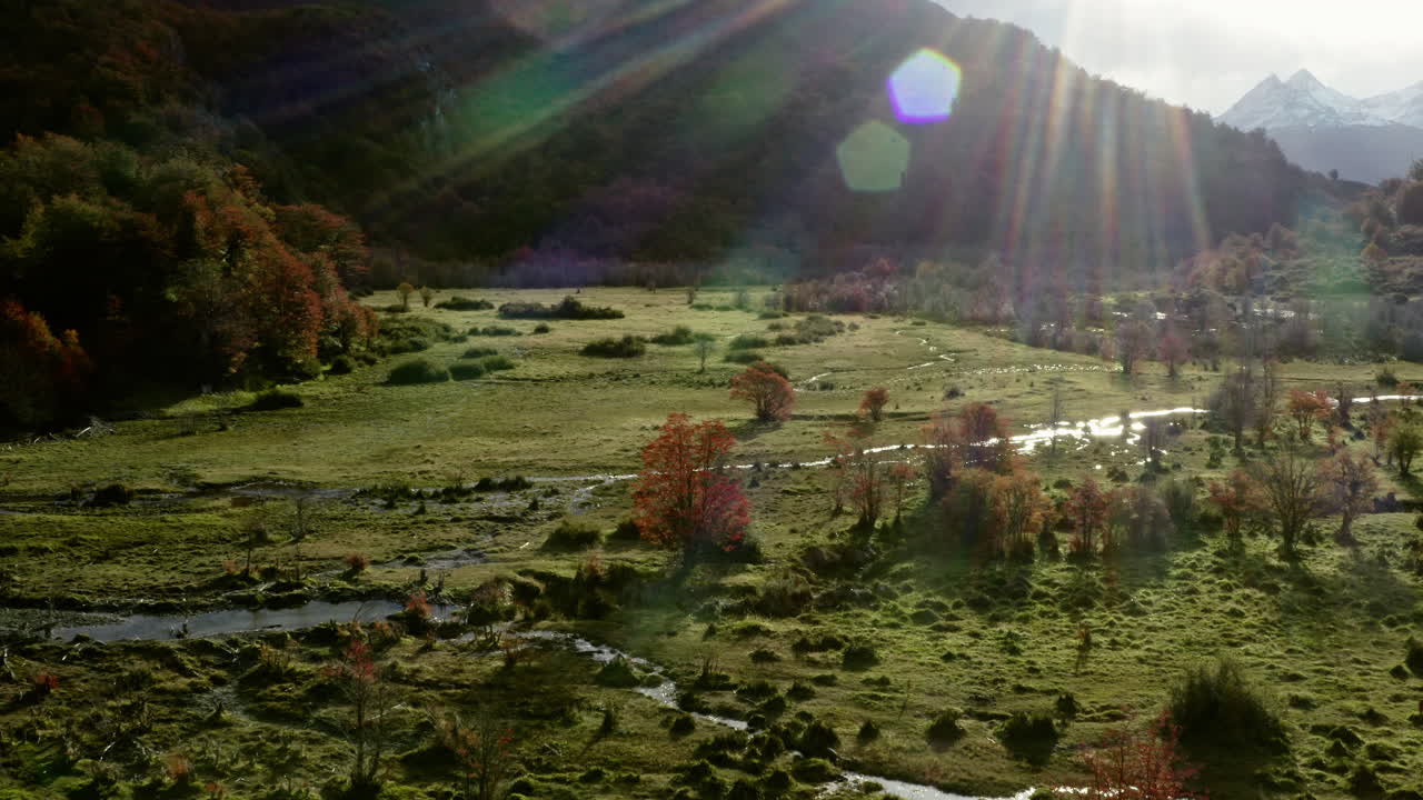 Sunbeam breaks through clouds over green pasture and slopes in remote Patagonian valley, aerial dolly over wetlands