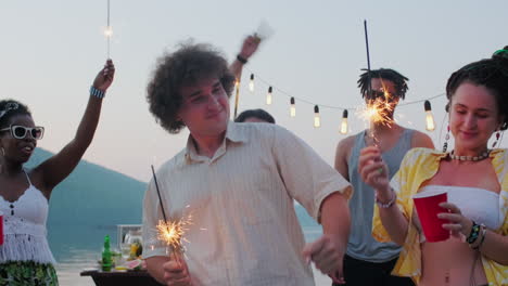 excited friends dancing with sparklers at lake party