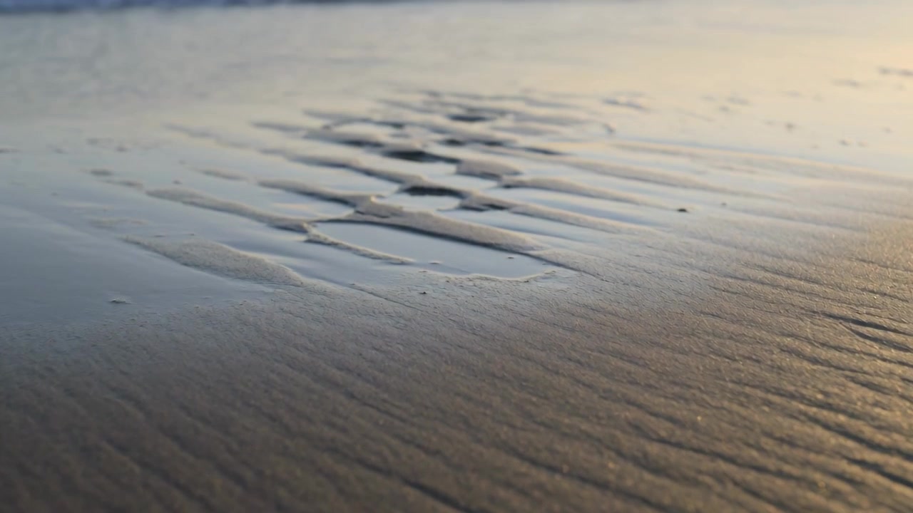 Advancing thin foamy wave pushing onto wet rippled sand during low tide, carving narrow tidal pools