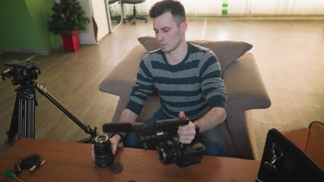 young man in striped sweater holding camera lens, preparing for filming, organizing photography equipment, working on table in modern home studio with professional tools and gadgets