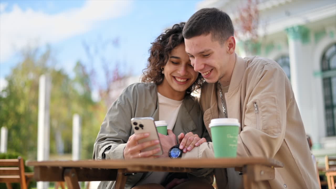 A happy couple outdoors near a cafe. Looking in the phone, laughing, talking, coffee. Autumn atmosphere. Slow motion