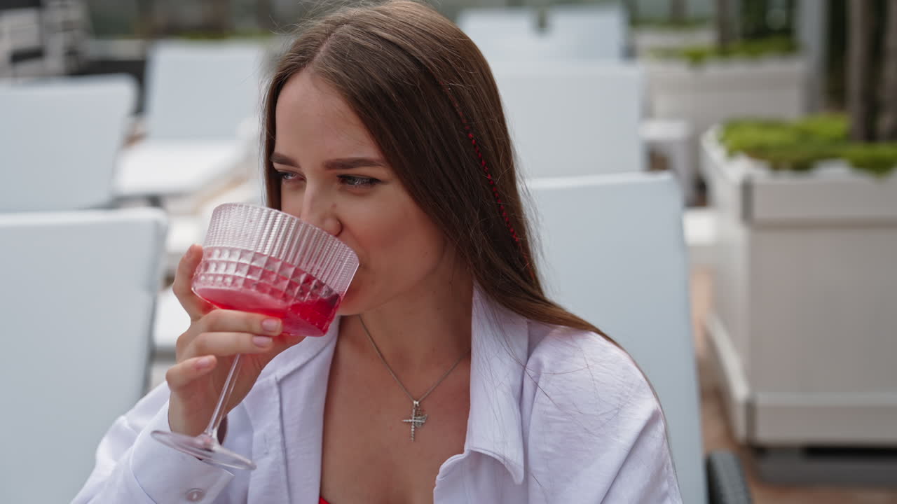 Attractive young girl smiling and taking sip from a wide wineglass. Lady sitting in a deckchair and relaxing with a cocktail. Blurred backdrop.