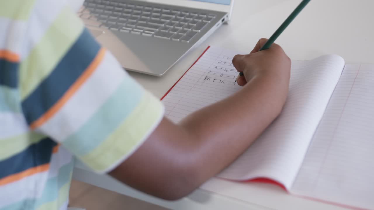 Un bambino che posa la matita e poi scrive calcoli matematici su un quaderno a righe a un tavolo bianco, facendo i compiti.
