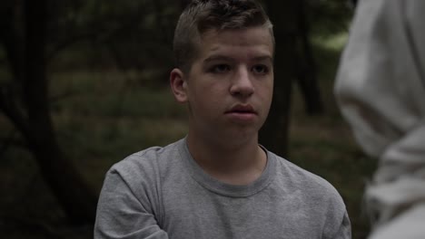 a teen boy stands to his feet and looks at man wearing robe in forest