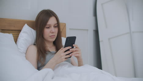 woman in morning sitting in bedroom leaning against the soft leather back of bed. write messages with your smartphone use apps to information