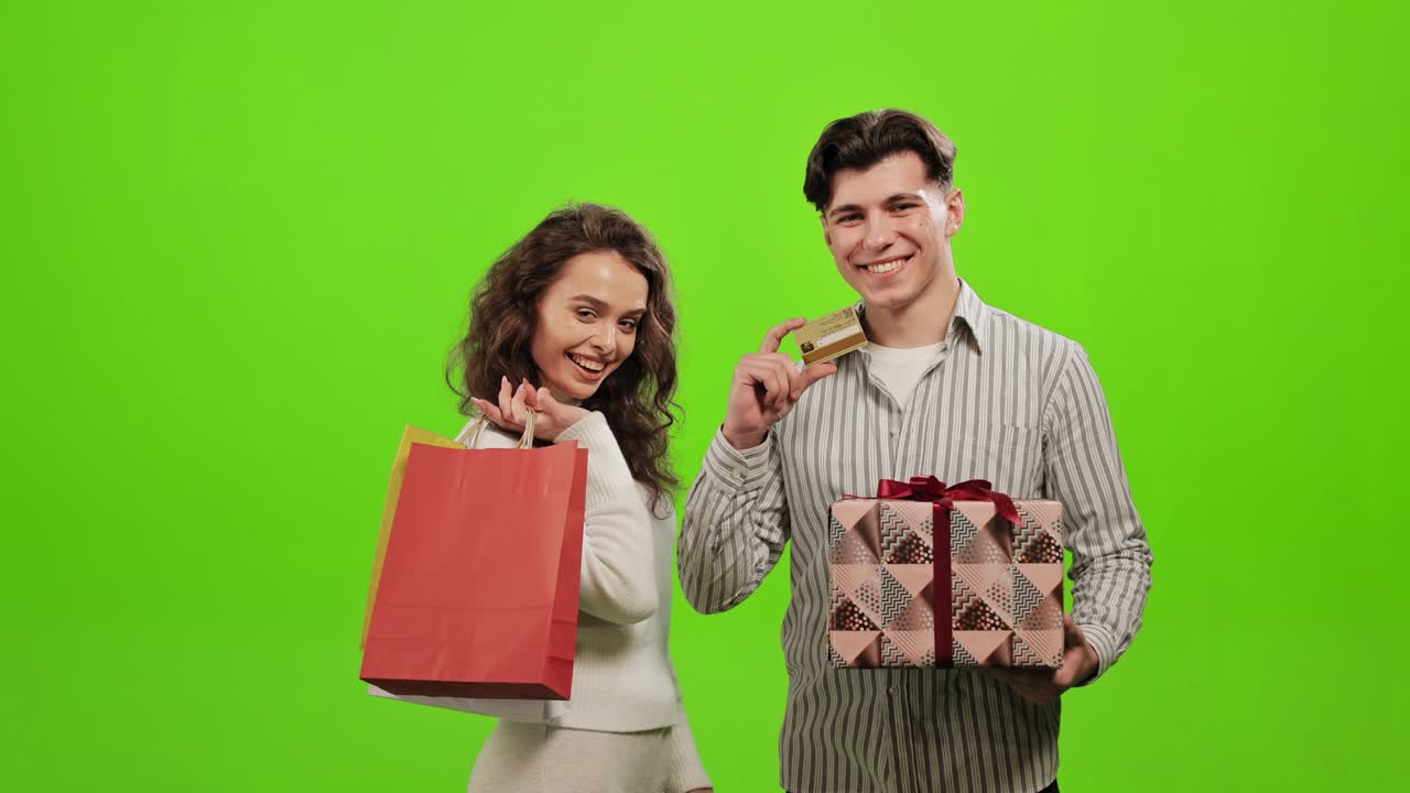한 남녀가 서서 카메라를 바라보고, 한 여인이 쇼핑백을 들고, 한 남자가 선물과 은행카드를 들고, 웃고, 초록색 바탕에 서 있다.