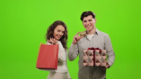 un hombre y una mujer están de pie y mirando a la cámara. una mujer tiene bolsas de compras. un hombre tiene un regalo y una tarjeta bancaria. están sonriendo. están de pie sobre un fondo verde. pantalla verde. 4k.