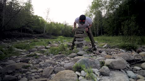 frustrated male hiker balances large pile of jagged stones with scattered rocks and dark forest background