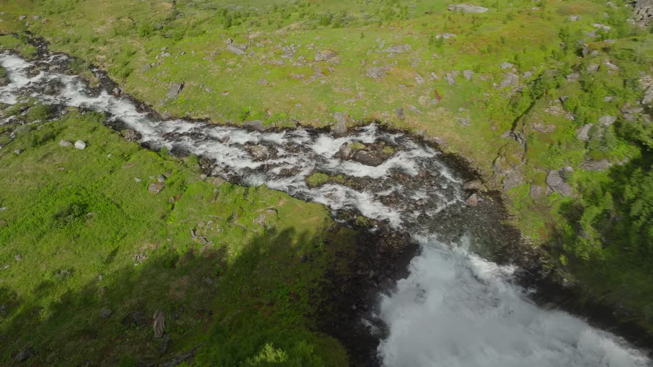 Filmato aereo con drone che segue una piccola cascata che scende da una collina in un fiume, circondata da pascoli verdi e vivaci e un paesaggio rurale sereno alla luce naturale del giorno