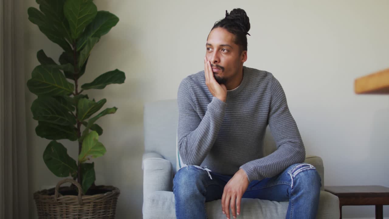 Thoughtful biracial man with dreadlocks in hair bun sitting in armchair in living room looking away