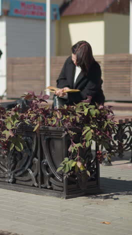 chica blanca sentada en un espacio público hojeando páginas de un libro pensativo, con el fondo mostrando una olla de flores balanceándose en el viento, peatones caminando y una máquina expendedora cerca