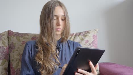 Portrait-of-young-blonde-girl-sitting-on-sofa-concentrating-on-writing-on-graphic-tablet