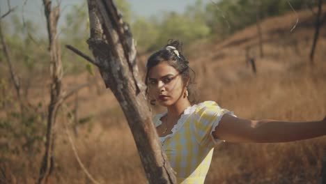 dreamy 35mm film look of young, attractive indian woman standing behind a tree in a sunny rural setting, looking flirtatiously at the camera before glancing downward, shallow depth of field