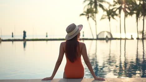 Vista-Trasera-De-Una-Modelo-Femenina-En-Forma-Que-Lleva-Un-Traje-De-Baño-Monokini-Naranja-Y-Un-Sombrero-Se-Sienta-En-El-Borde-De-La-Piscina-Apoyándose-En-Los-Brazos-Al-Atardecer